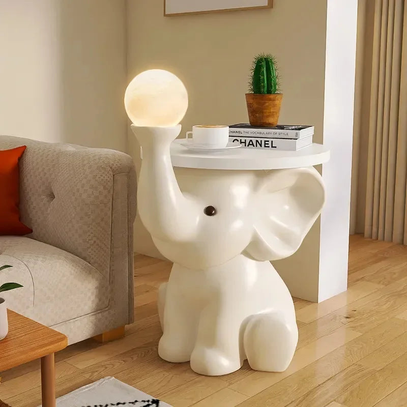 A white Elephant Bedside Table holding a glowing orb lamp and a stack of books with a small potted cactus on top, in a cozy bedroom setting.