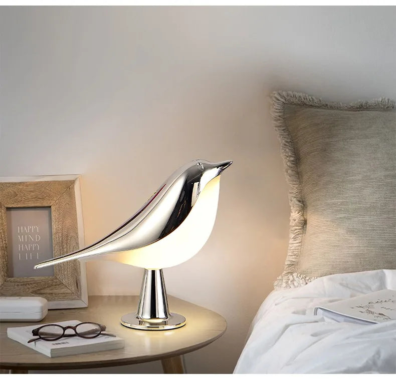 A Bird Table Lamp sits on a nightstand beside a bed with a textured pillow. The illuminated lamp brightens up a pair of glasses and a picture frame, and includes an easy-to-use touch sensor for convenience.