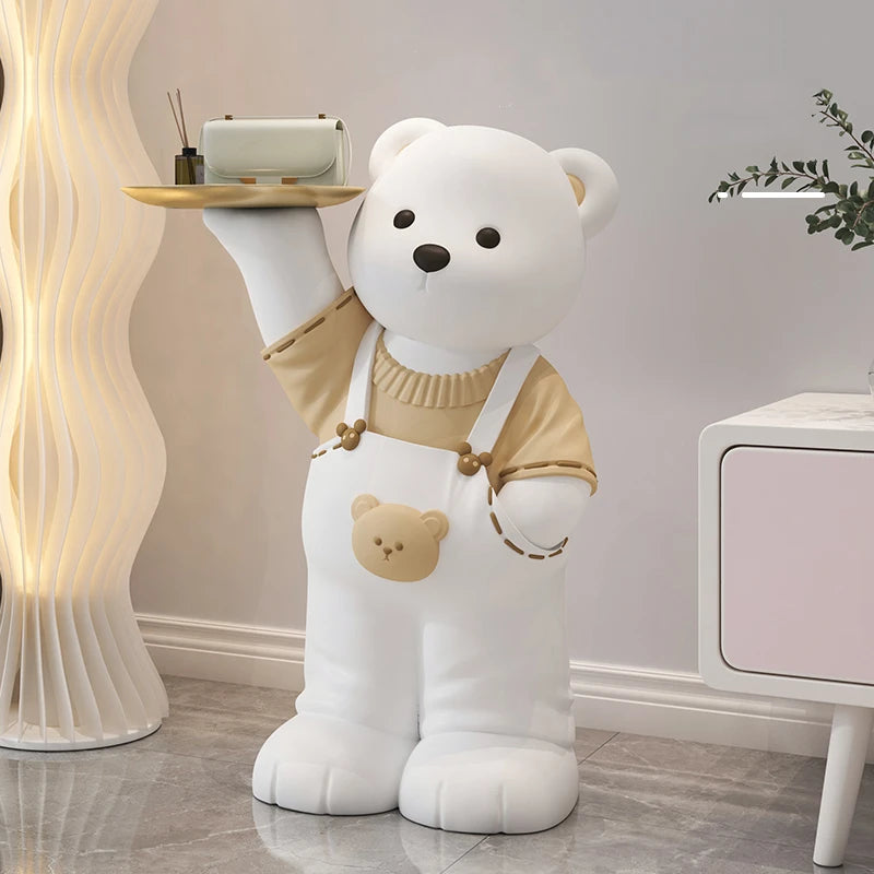 Life-sized bear art piece in a sweater and overalls, holding a tray with a phone, in a modern room with a wavy floor lamp.