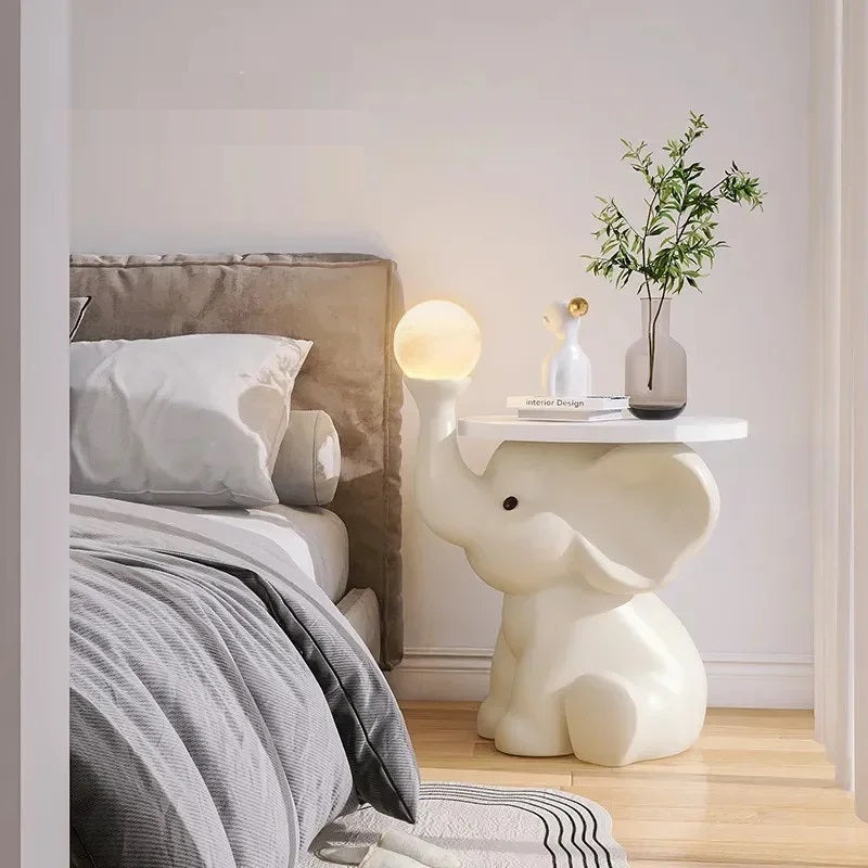A charming bedroom with an Elephant Bedside Table holding a lamp, vase with greenery, and a small figurine.