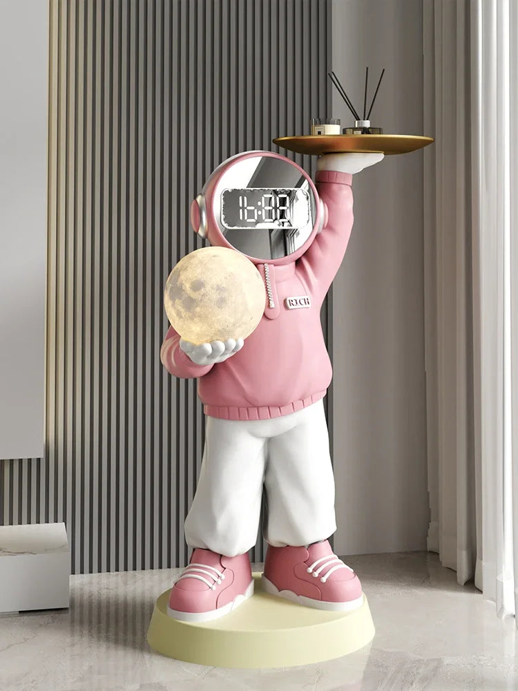 A whimsical sculpture of an astronaut in a pink hoodie holding a glowing moon in one hand and a tray with a cup and saucer on the other, with an astronaut-themed clock face for a head