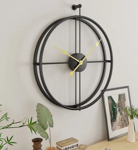 Large minimalist ring wall clock, featuring yellow hands against a black backdrop, mounted on a light-colored wall beside framed artwork and decorative plants.