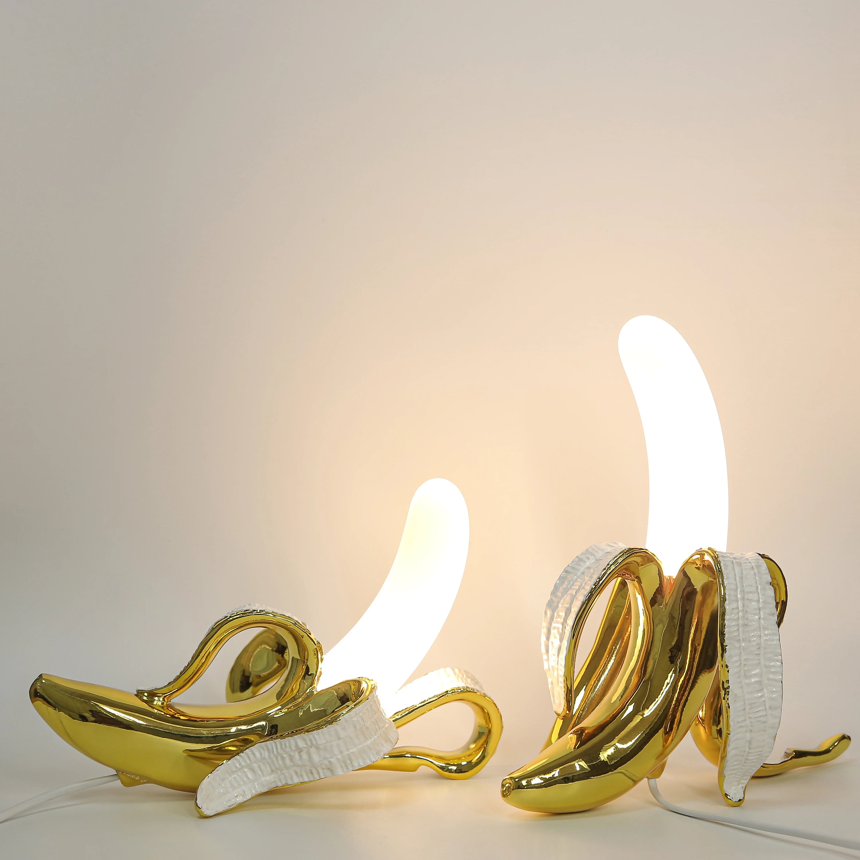 Two golden Banana Table Lamps with illuminated white bulbs, set against a plain light background.