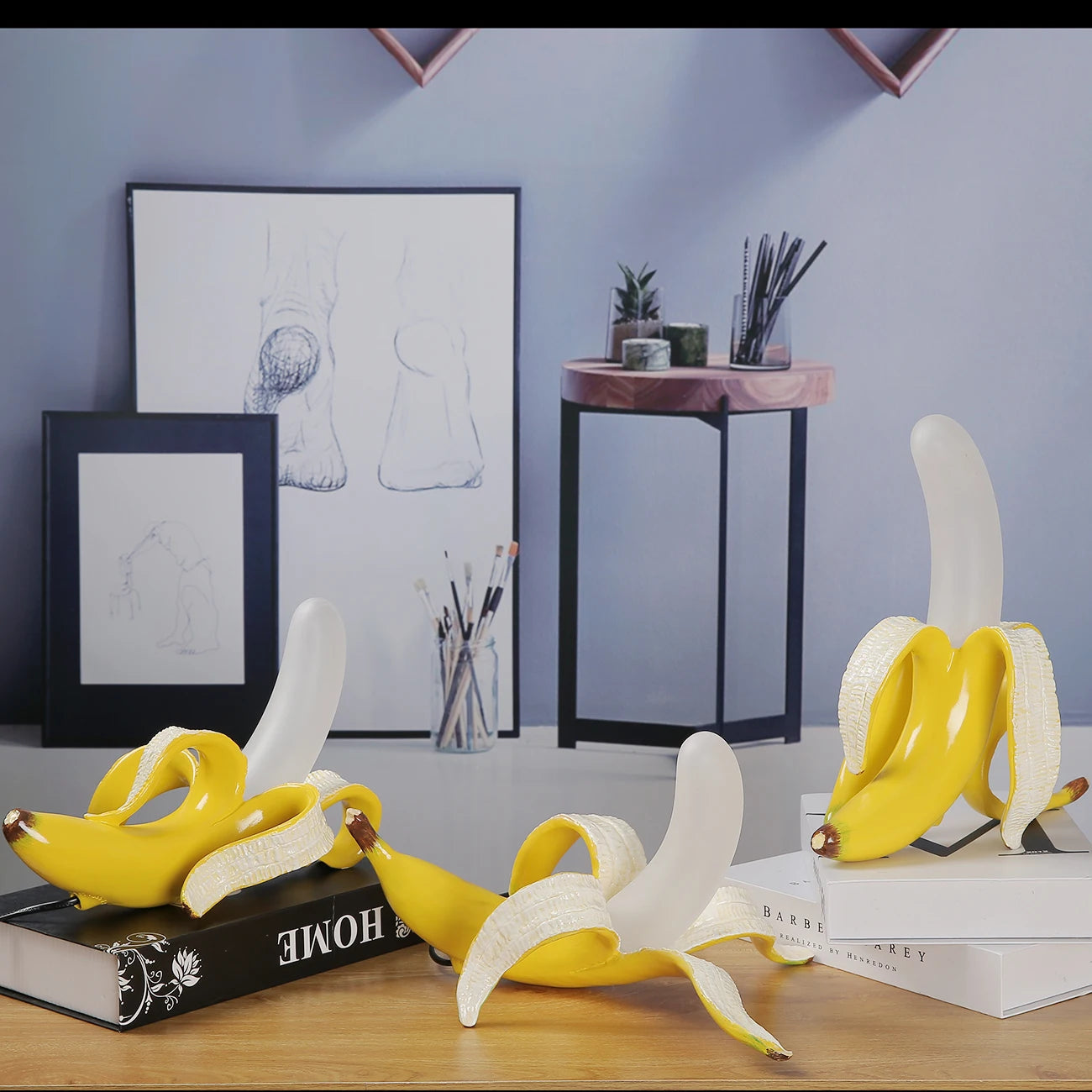 A creative display featuring a unique Banana Table Lamp over books on a desk, with framed sketches and plants in the background.