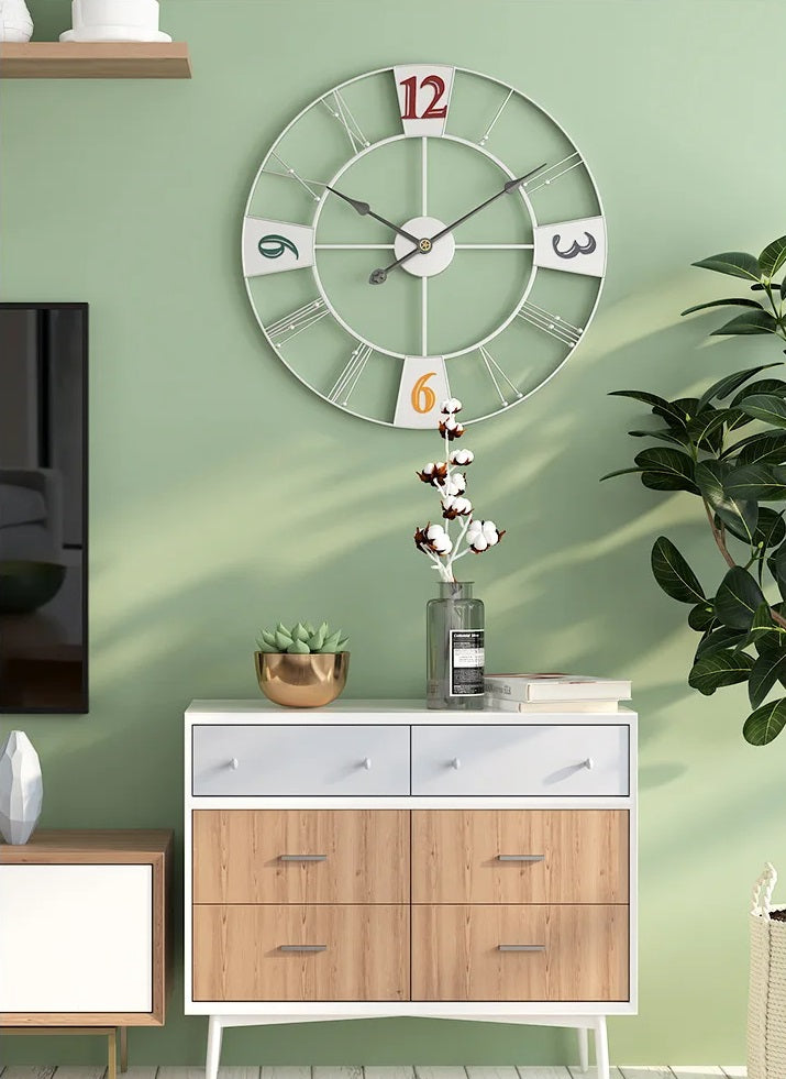 A large wall clock with Roman numeral cutouts above a modern wooden cabinet with decorative items and plants against a green wall.