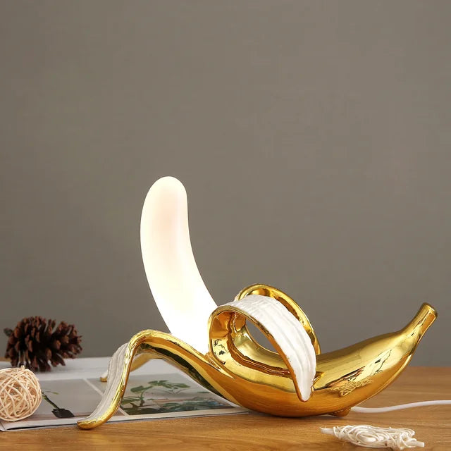 A golden lamp designed like a genie's lamp, with a glowing white light emerging from the spout, displayed on a wooden table against a gray background. This unique banana table lamp creates a cozy atmosphere.