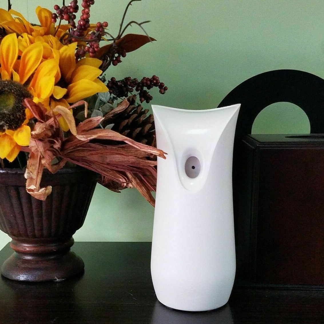 A modern white SpyCam air freshener dispenser stands between a vase with artificial flowers and a dark brown wooden box on a table with a teal wall in the background.