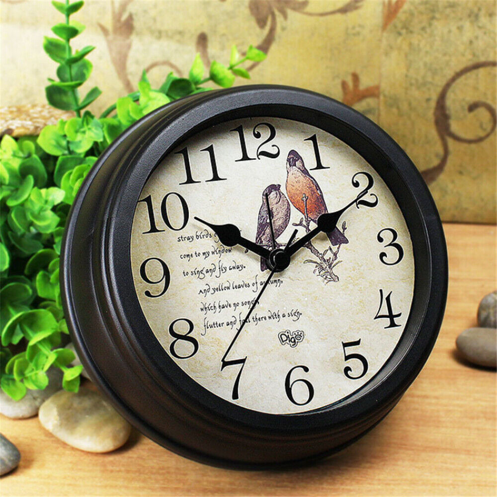Analog wall clock with a vintage bird illustration on its face, displaying the time approximately at ten minutes past ten, placed on a wooden surface with greenery and pebbles around it.