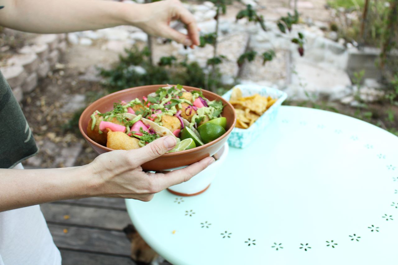 A person out of frame holds a terracotta bowl holding three halibut cheek tacos garnished with avocado crema and cilantro, and several lime halves. The other hand is sprinkling additional cilantro leaves on the dish. In the background out of focus is a table with a dish holding chips and some foliage in the background.