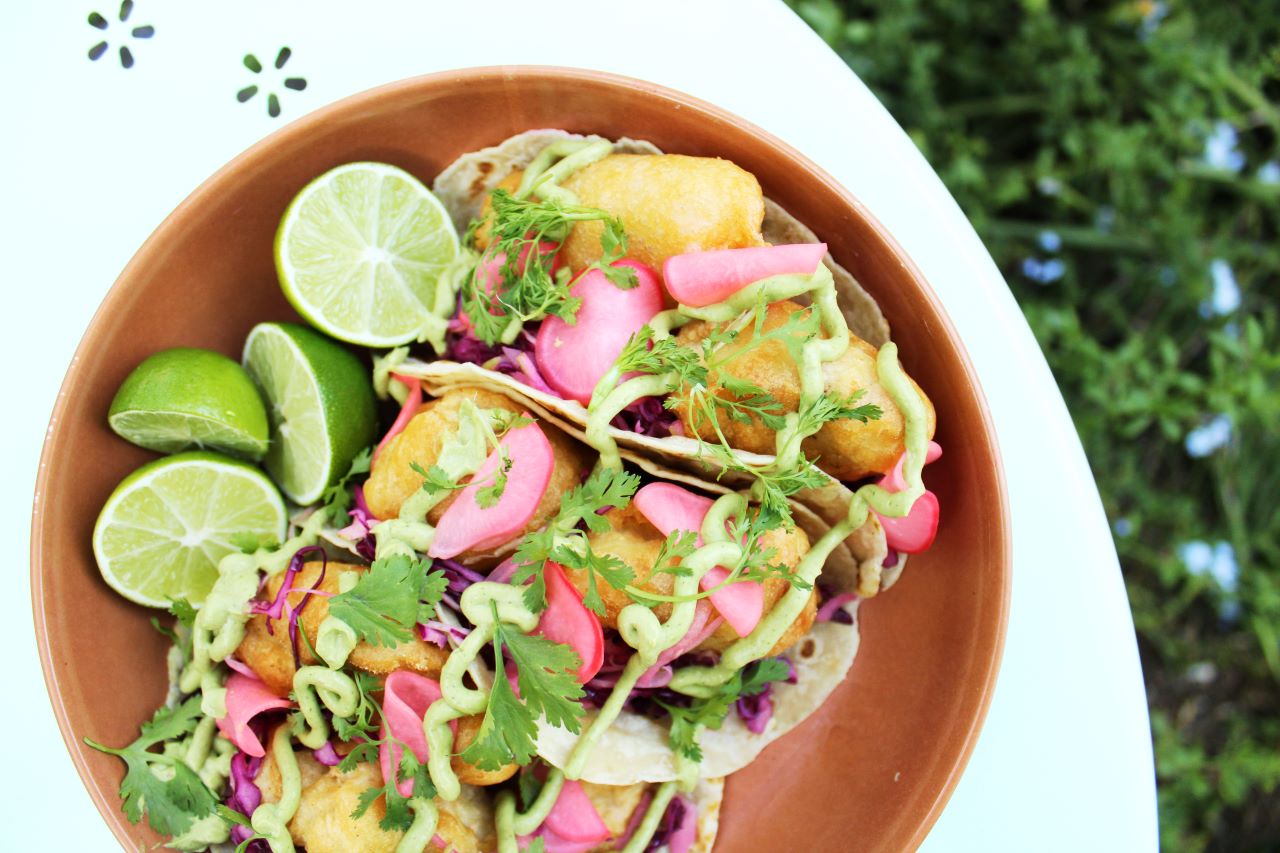 Three tortillas filled with beer battered halibut cheeks and red cabbage slaw are topped with a green avocado crema and pickled radish slices. They are siting in a terracotta bowl next to several halved limes. A white table and greenery makes up the background. 
