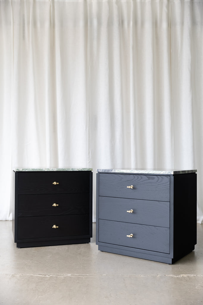Pair of black wooden bedside cabinets with 3 drawers each, brass T-shaped handles and marble tops