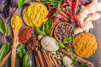 Shot from above of many spices on a wood table — ground spices in wooden spoons and whole spice ingredients. Turmeric, mustard, garlic powder, whole cinnamon, ginger, anise, nutmeg, and more.