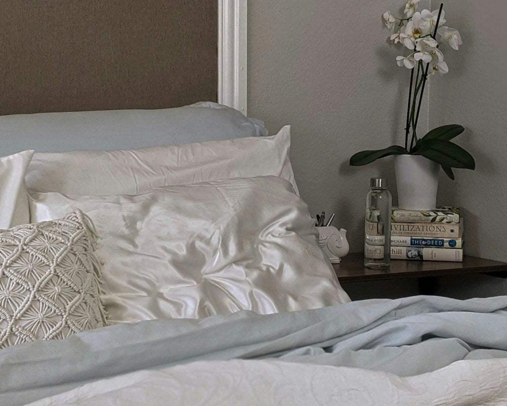 Detail of a bed with white and light blue bed linen, a white silk cushion next to a brown bedside table with a white orchid, books and a bottle of water.