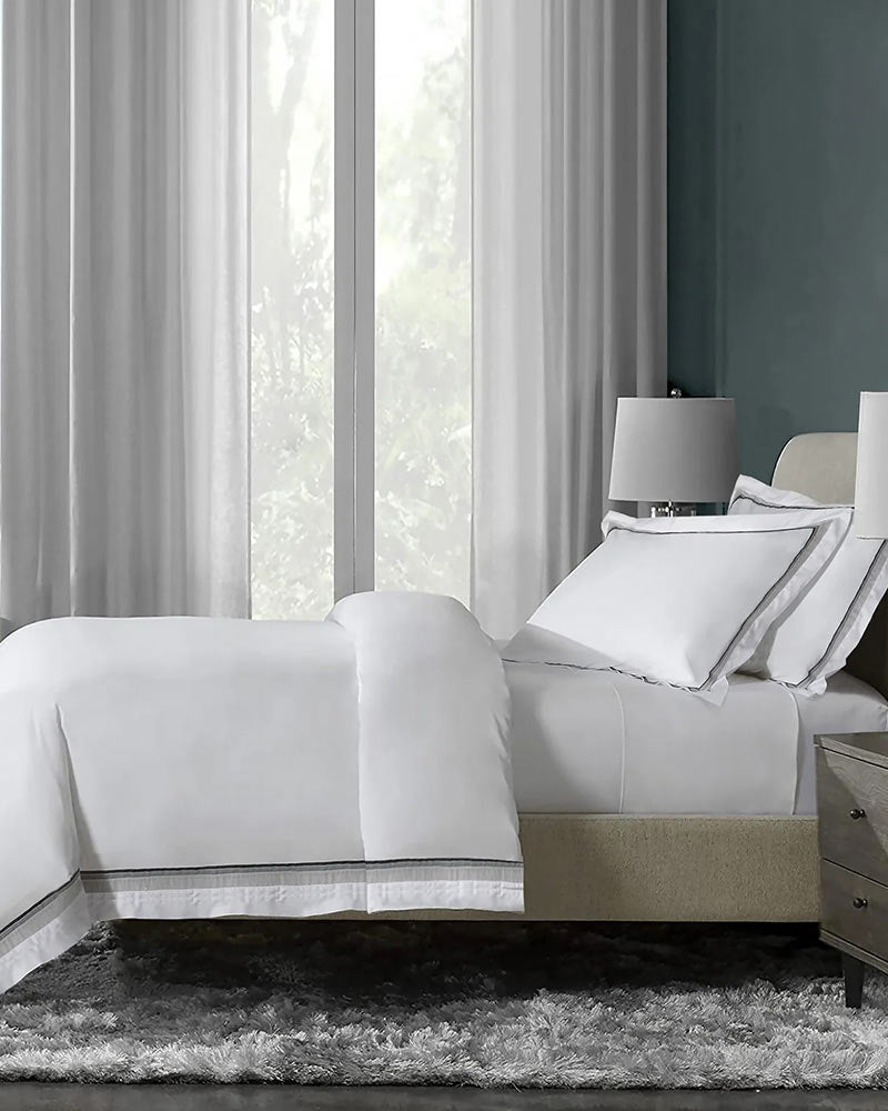 White duvet cover set with a comforter cover, a grey stripe on its edges, and two pillowcases on a bed next to two side tables with a lamp.