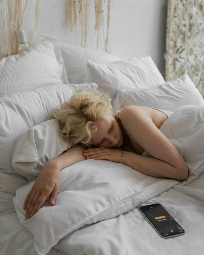 A blonde woman with short hair, lying on a bed with white bedding next to a cell phone with an alarm clock.