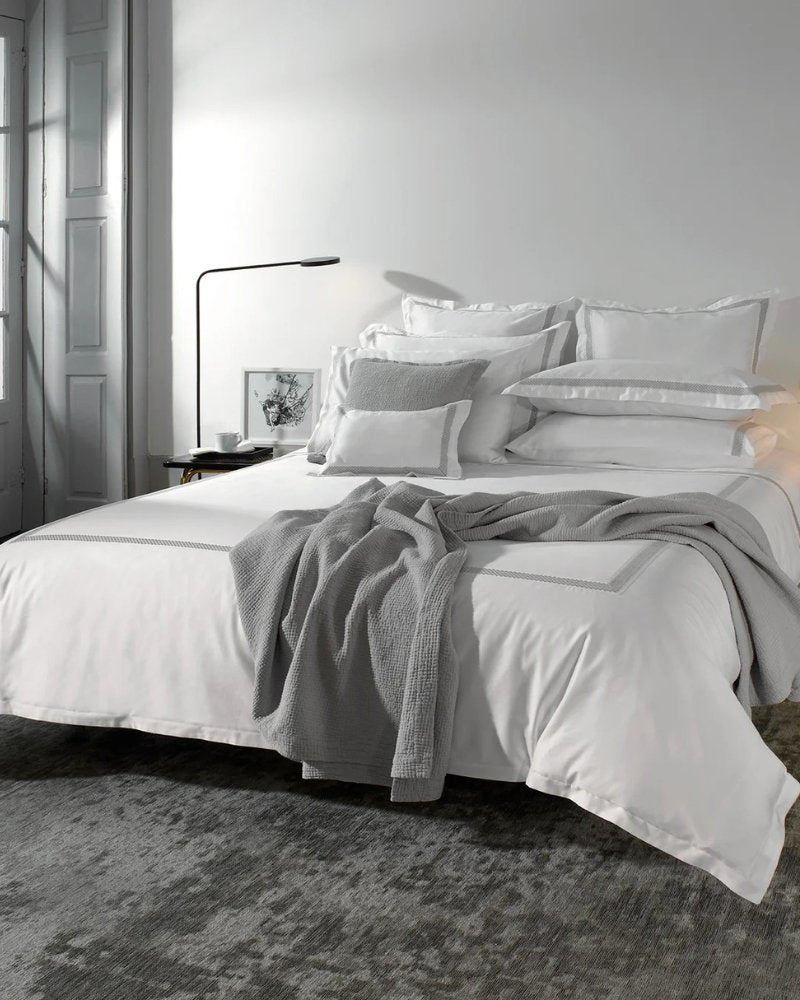 Bedroom with white walls, a high window, and a bed with white bedding from Amalia Home Collection next to two side tables decorated with lamps and books.