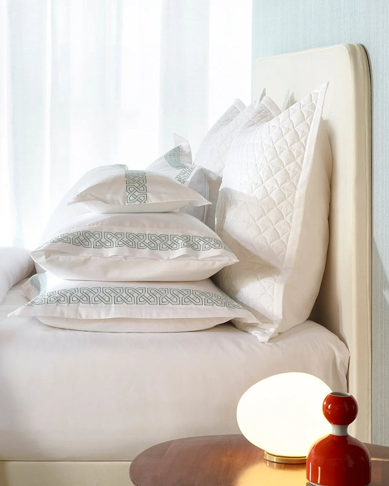 Blue bedroom wall and a headboard, a lamp, and a stack of pillows on top of the bed with daylight coming through the window curtain