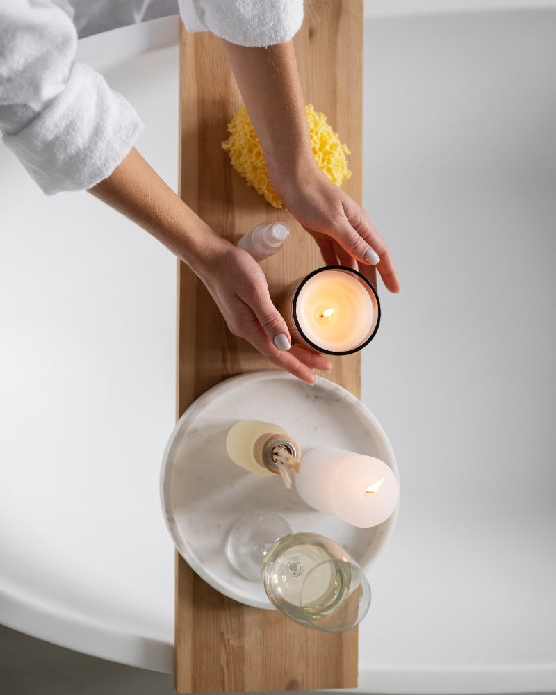 Woman placing lighted candles, a cup of wine, and a reed diffuser on top of a bath wood board.