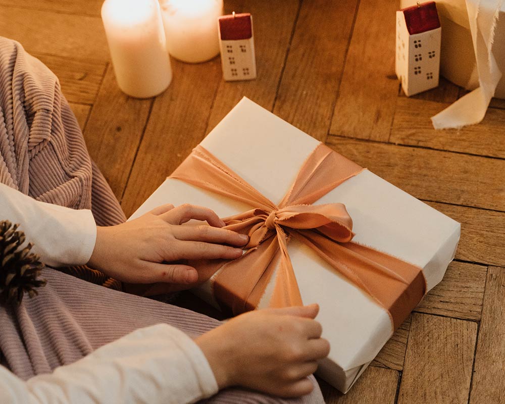 A person sitting on the floor with lit candles unwrapping a present with white wrapping paper and an orange bow.