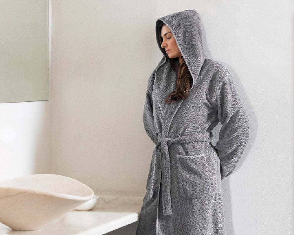 Brown-haired woman wearing a grey bee waffle hooded bathrobe from Graccioza next to a sink in the bathroom.