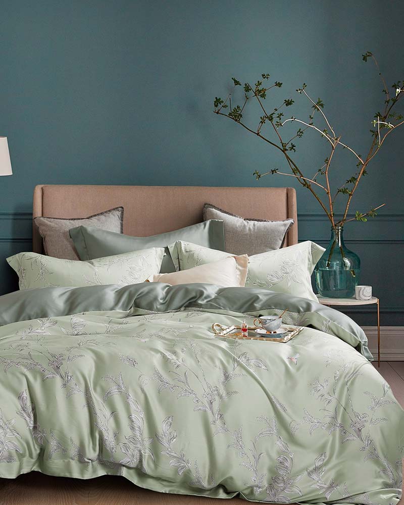 Floral green Micromodal bedding from St. Pierre in a bed with a fabric headboard next to a bedside table with a plant in a green wallpapered bedroom.