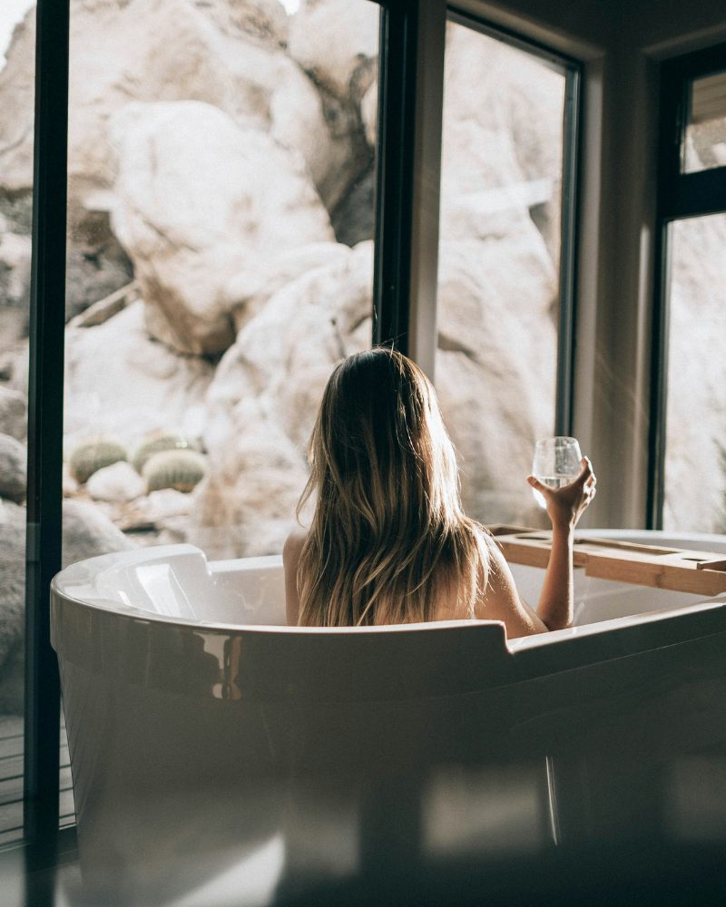Blonde woman taking a relaxing bath while drinking a glass of wine in a bathroom with Nature view.