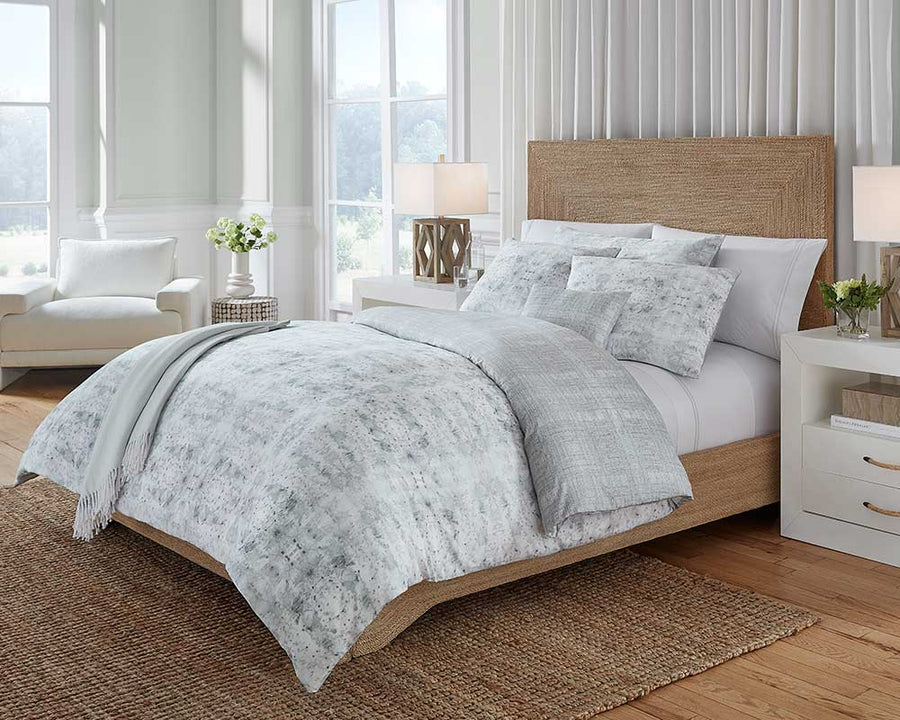 Grey bedding from Sferra in a bedroom next to two big windows, a wooden floor, and white furniture.