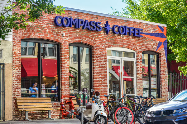 Exterior view of Compass Coffee in Washington DC, showcasing its vintage brick design, prominent brand signage, and outdoor seating area. The bustling urban location is evident with bikes and scooters parked outside, inviting locals and visitors for a cup of Real Good Coffee.