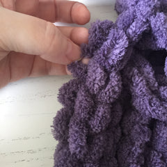 Purple chenille loop yarn showing a horizontal chain stitch, held vertically. A hand pulls on the stationary loop under the chain.