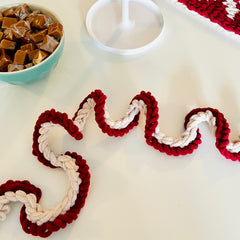 A red and white chenille yarn garland on a white surface with a bowl of candied caramels in the top left corner.