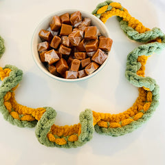 A green and yellow chenille yarn garland on a white surface with a bowl of caramel candies.