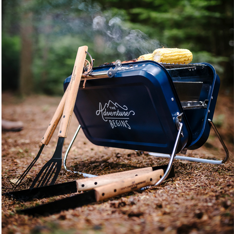 portable bbq grill alongside wooden grill tools in a campsite with corn on the grill