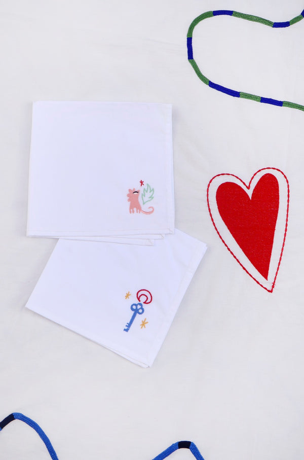 The first napkin has a pink lion with a green leaf and small red heart embroidered in the corner, and the other has a blue key with stars and a red moon outline.  | © Pangea 2024