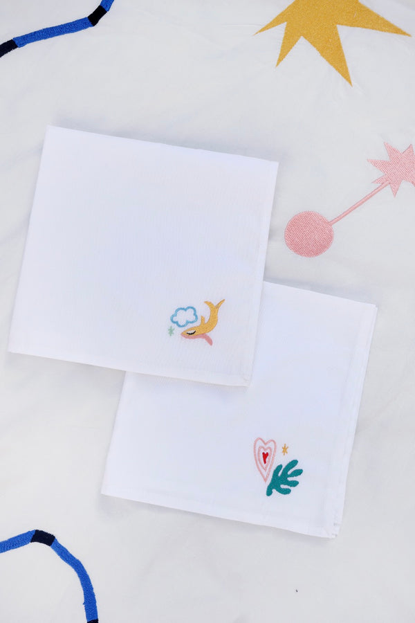 The first napkin has a yellow fish with the blue outline of a cloud, and the second has a pink and red heart and green filled in leaf embroidered. | © Pangea 2024