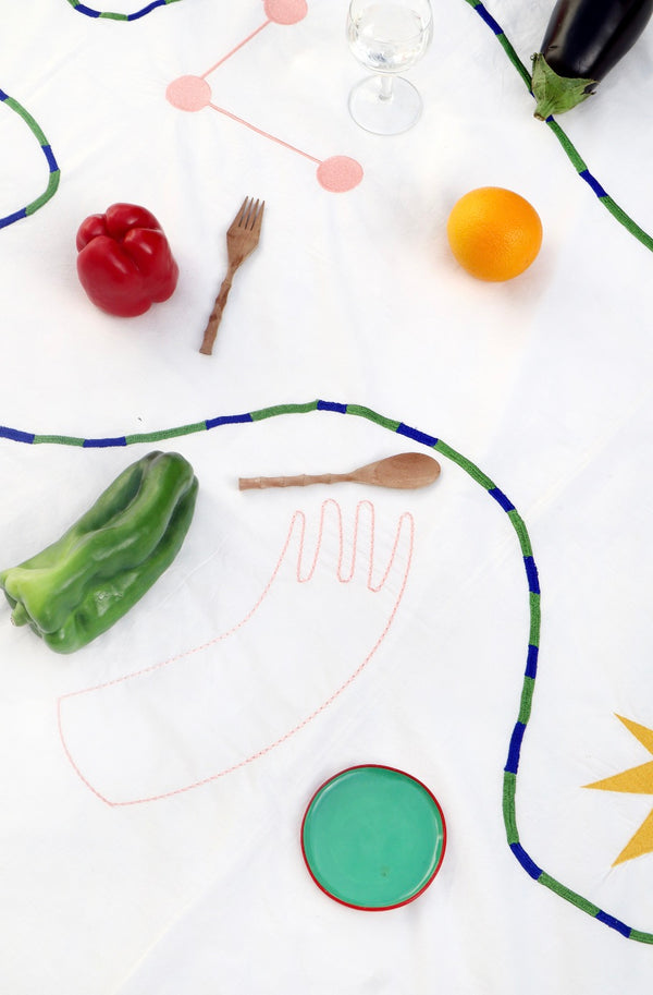 A close up image of the tablecloth, showing the green and blue embroidered swirl, and the pale pink hand.  | © Pangea 2024