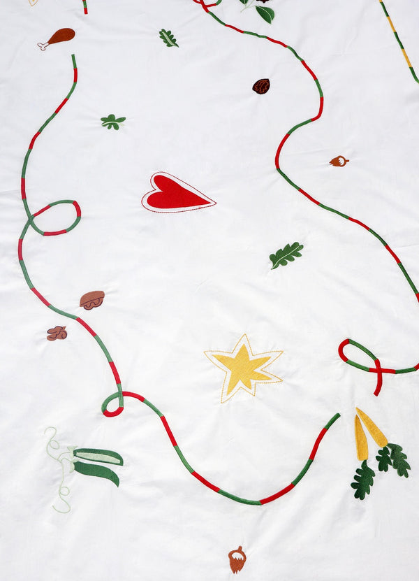 The white tablecloth has Christmas themed items embroidered on it: red heart, green leaves, green beans, carrots, chicken/turkey drumstick, a bright star and a red and green swirl covering the entire middle section. | © Pangea 2024