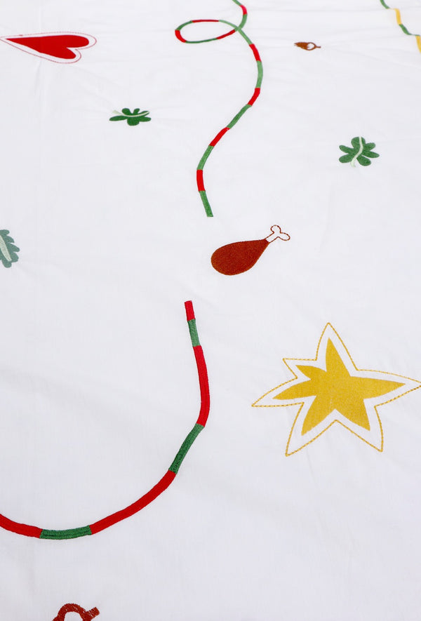 The white tablecloth has Christmas themed items embroidered on it: red heart, green leaves, green beans, carrots, chicken/turkey drumstick, a bright star and a red and green swirl covering the entire middle section. | © Pangea 2024