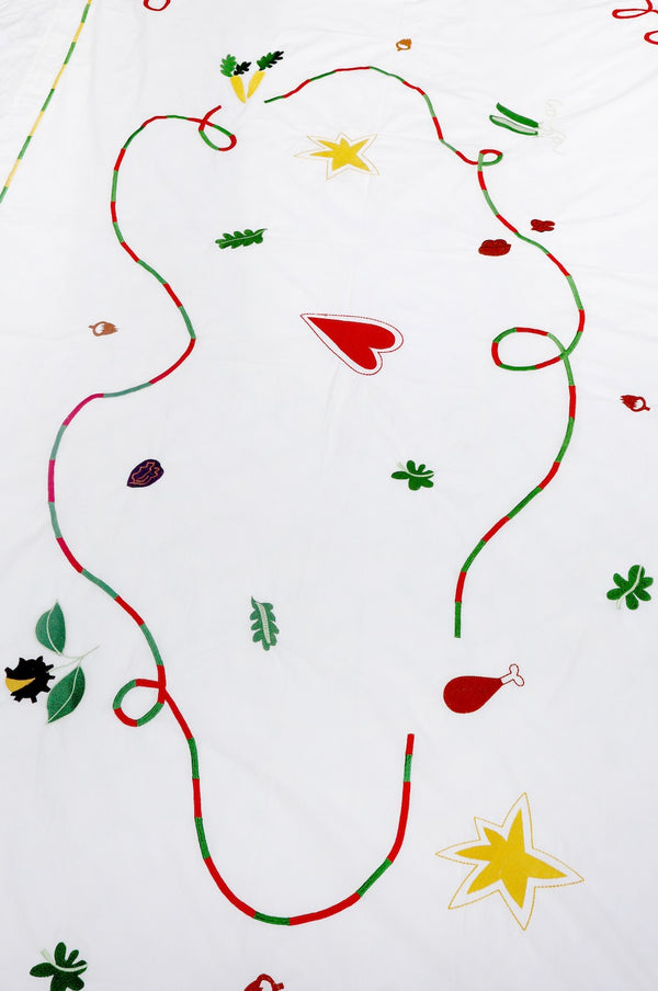The white tablecloth has Christmas themed items embroidered on it: red heart, green leaves, green beans, carrots, chesnuts, chicken/turkey drumstick, a bright star and a red and green swirl covering the entire middle section. | © Pangea 2024