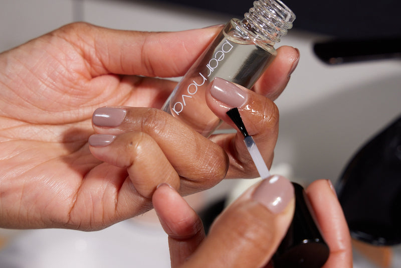 brown skin model with natural nails painted taupe holding a bottle of cuticle oil and applying it