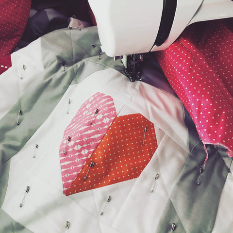 Close up of a quilt in progress. The quilt features a pink and orange heart inside a white chat bubble on a grey background. There are safety pins all over the quilt, and it is being fed through a Janome sewing machine.
