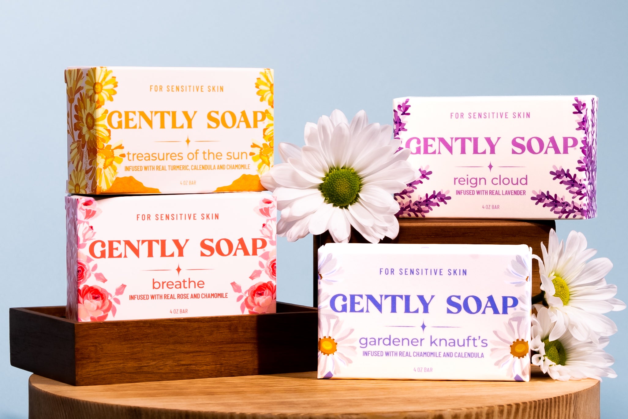 Four colorful bars of soap with floral designs displayed with daisies on wooden steps against a blue background.