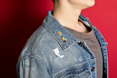 Person wearing a denim jacket with 2 inclusive pride flag enamel pins in the design of a heart and a peace symbol