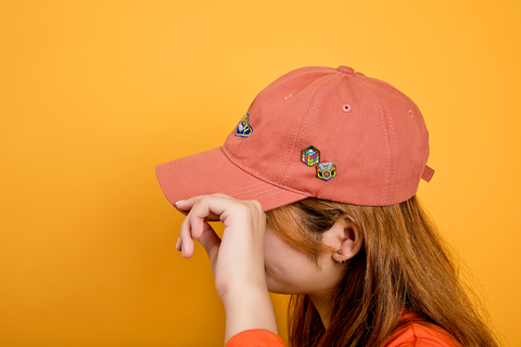 Person wearing an orange cap with 2 progress pride flag enamel pins in a design of a medal and a rubik's cube
