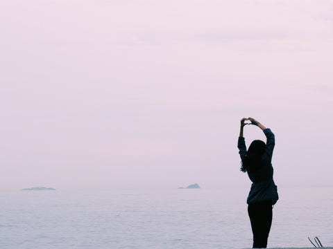 Woman in front of ocean at sunset holding up a heart symbol with her hands above her head.