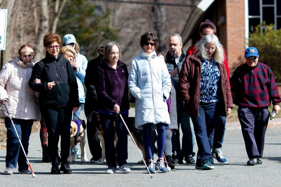 Ten people, both sighted guides and VLANJ program participants walking outside on a sunny day. Some have their guide dogs with them, and others have white canes. They are smiling and talking. 