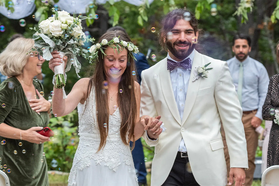 Anthony and Kelly walk down the aisle as newlyweds. Anthony wears a cream white tux and purple bow tie. Kelly wears a white dress with tactile details embroidered.