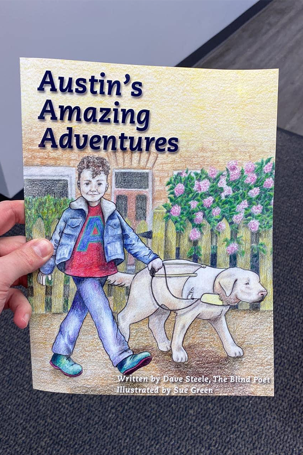 Filling the frame, a close up of Austin's Amazing Adventures. On the illustrated cover, Austin walks with his guide dog past wooden picket fences and a pink rose bush. Austin wears a blue jacket, a red top with a large letter 'A' in blue across the front, blue bottoms and green shoes.