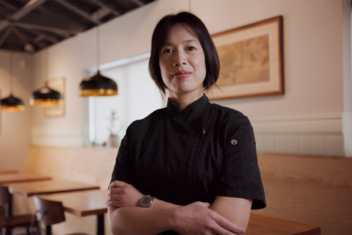 Headshot of Chef Christine Ha wearing a black chef jacket, standing in Xin Chao. The decor is light wood, earthy tones with simple black and gold hanging lights. On the walls, framed images can be seen.