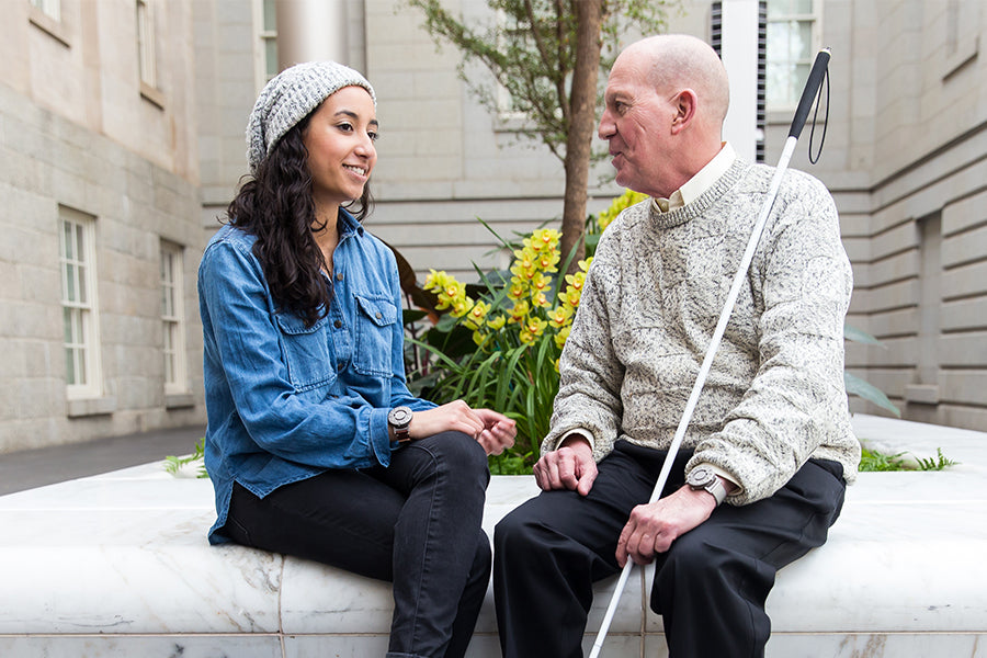A young lady and elderly gentleman are sat outside on a white marble bench surrounded by buildings. They are smiling at each other and in conversation. The lady wears a blue demin button shirt, black jeans, and the Eone Bradley Classic. The gentleman wears a grey heavy knit sweater, black suit pants and an Eone Bradley Silver Mesh. A long white cane can be seen resting against the shoulder of the gentleman.