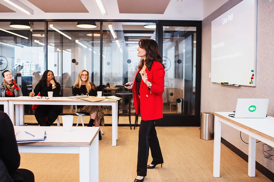 Taken from the side: Sharon faces a class of individuals, mid stride with her left hand raised to her side. she wears a red blazer and black suit pants. In the background, to the left, three participants can be seen smiling and engaged with Sharon.